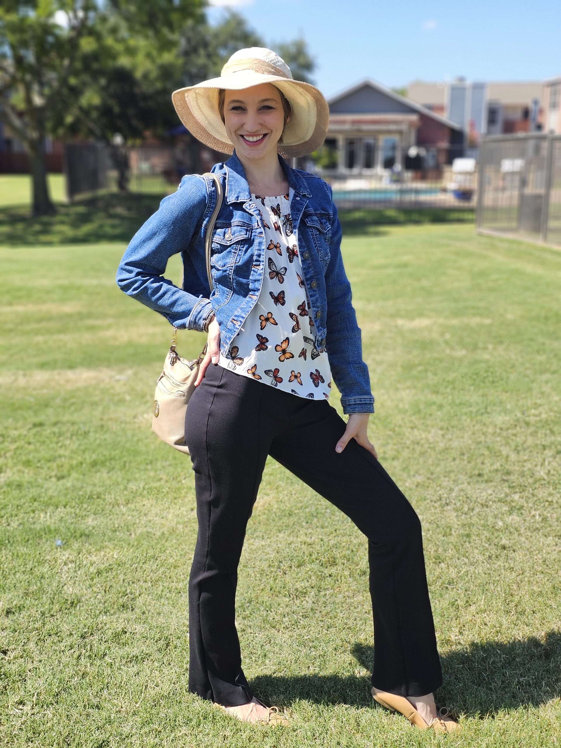 Yevgeniya Altamirano standing against a grassy green field with a building in the bakcground, wearing a summer hat, butterfly top, jean jacket, Spanx black pants, and tan ballet flats.