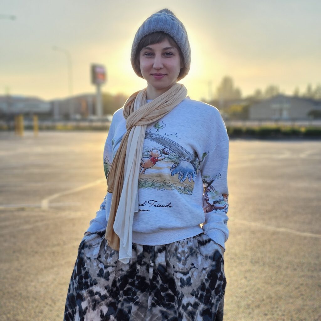 Yevgeniya Altamirano standing against a sunset wearing a winter hat, light scarf, and WInnie the Pooh sweater with a tie-dye skirt.