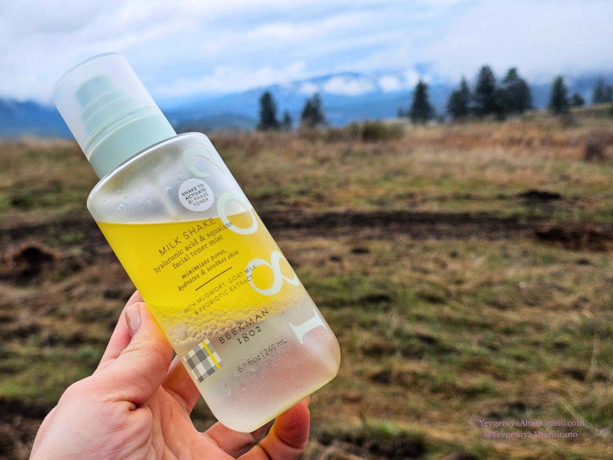 Female hand holding the regular size bottle of Beekman 1802 Milk Shake Toner against a sparce forest background.